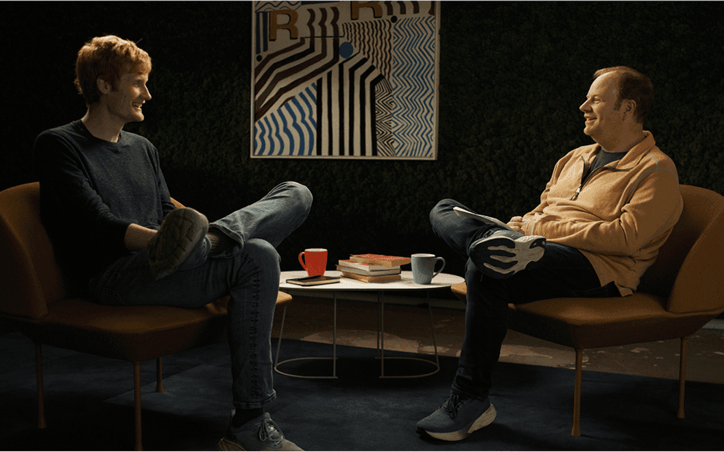 Image of Cursor CEO Michael Truell on the left and Rippling CEO Parker Conrad on the right. Both are seated across from eachother in a living room set and they are both talking candidly
