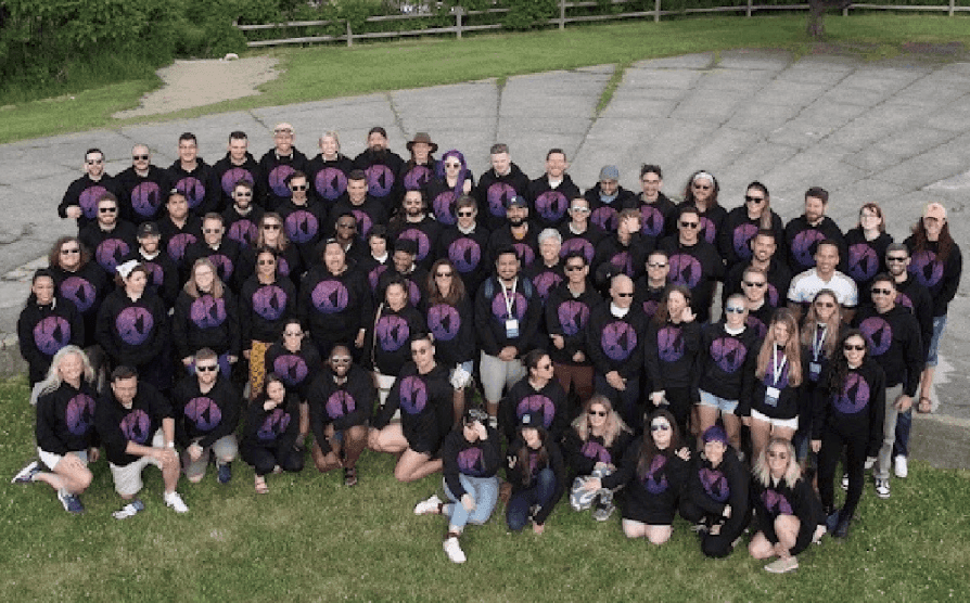 Large group gathered outdoors wearing matching black shirts with purple circular logos, posed together on grass.