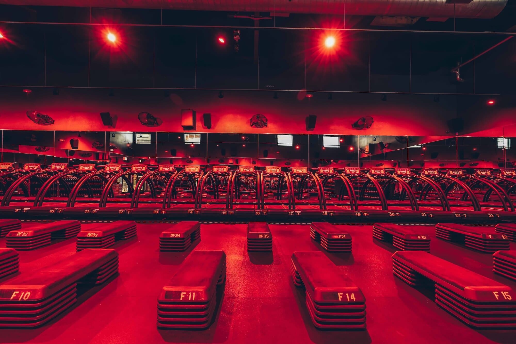 Rows of treadmills and step platforms in a dark fitness studio lit by red lights.