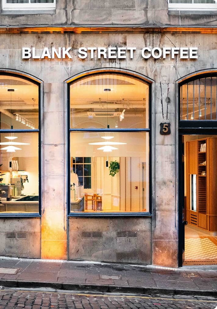 Exterior of Blank Street Coffee shop with large arched windows, warm interior lighting, and a stone facade on a cobblestone street.