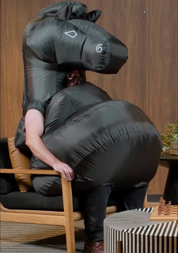 Person wearing a black inflatable horse costume sitting in a wooden chair next to a striped table with chess pieces.