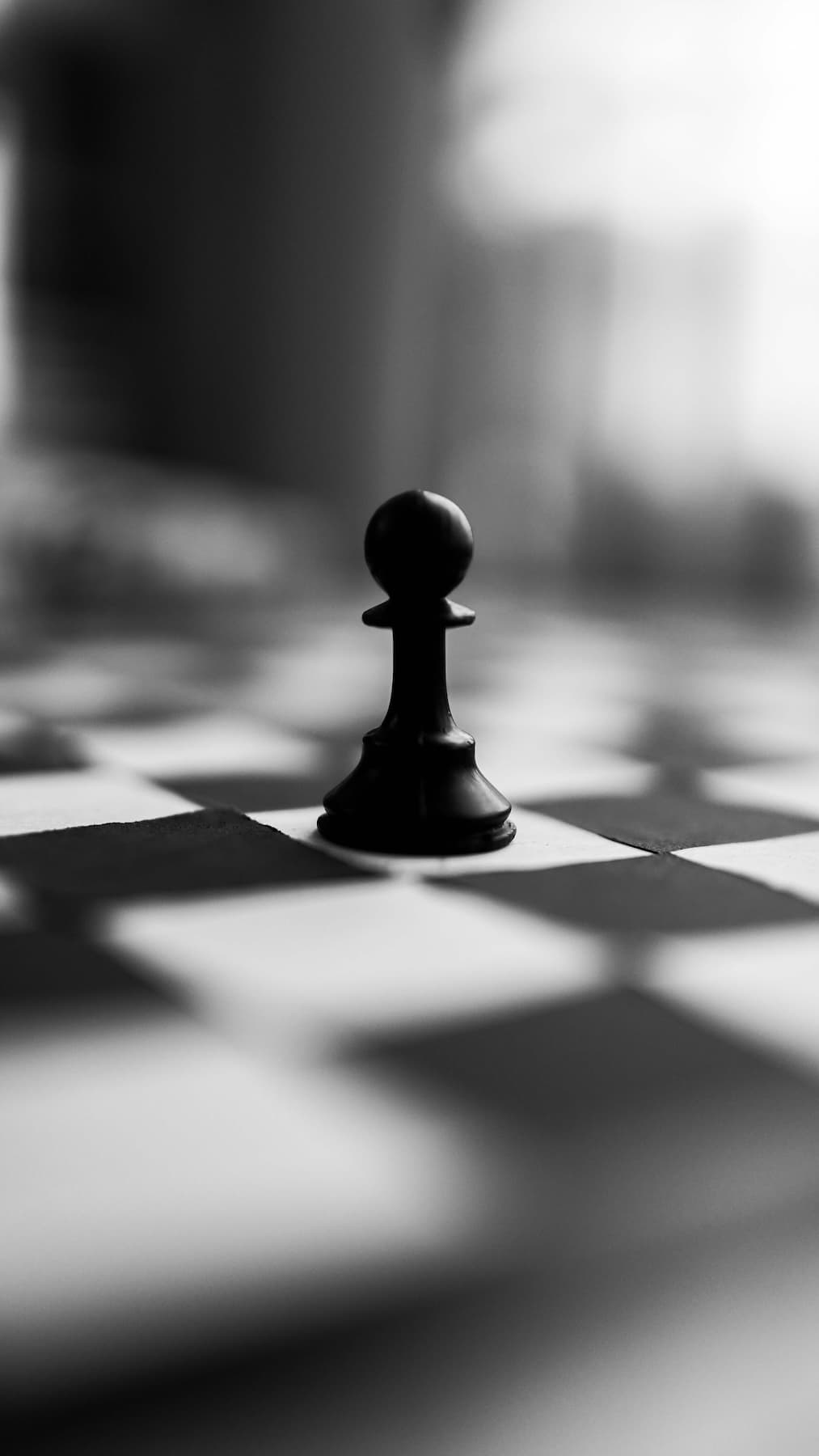 Black-and-white close-up of a lone pawn on a chessboard with blurred pieces behind it.