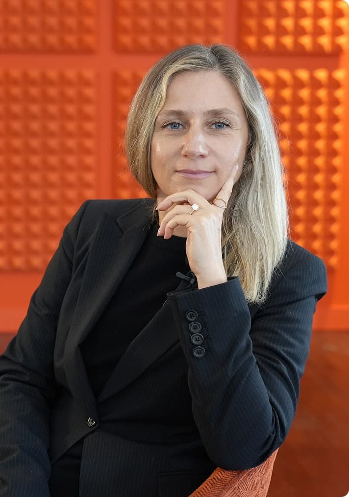 A woman in a black blazer poses thoughtfully with hand on chin against a bold orange textured wall background.